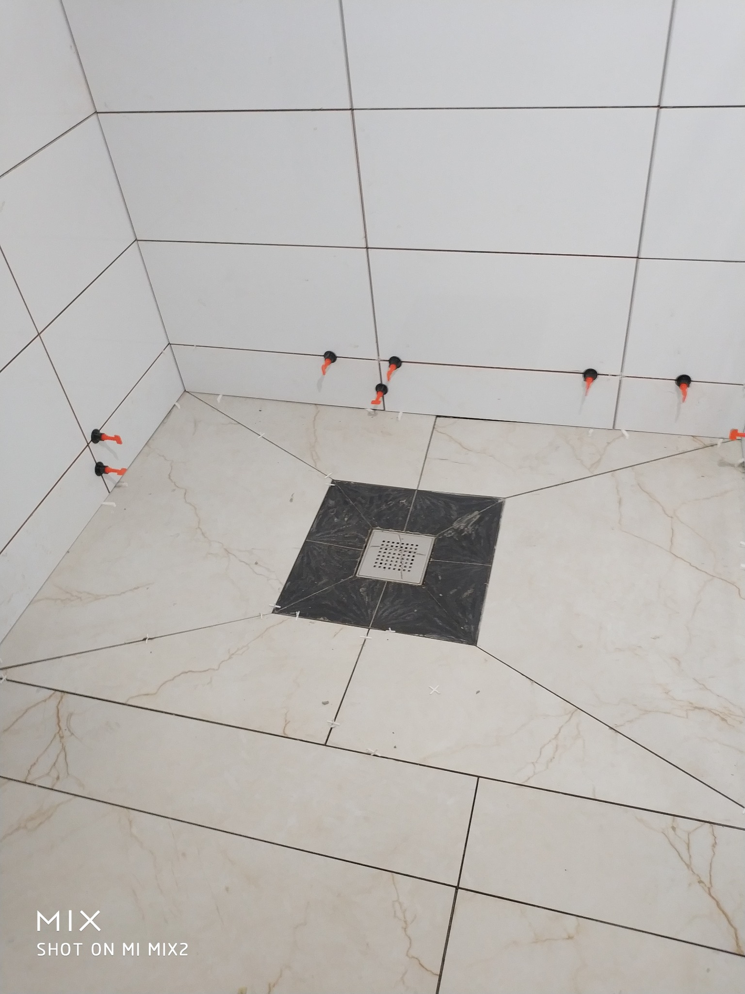 Układanie płytek w łazience, widoczne białe płytki ścienne oraz jasne płytki podłogowe z marmurkowym wzorem, system poziomowania płytek z pomarańczowymi klinami, czarny kwadrat wokół odpływu...