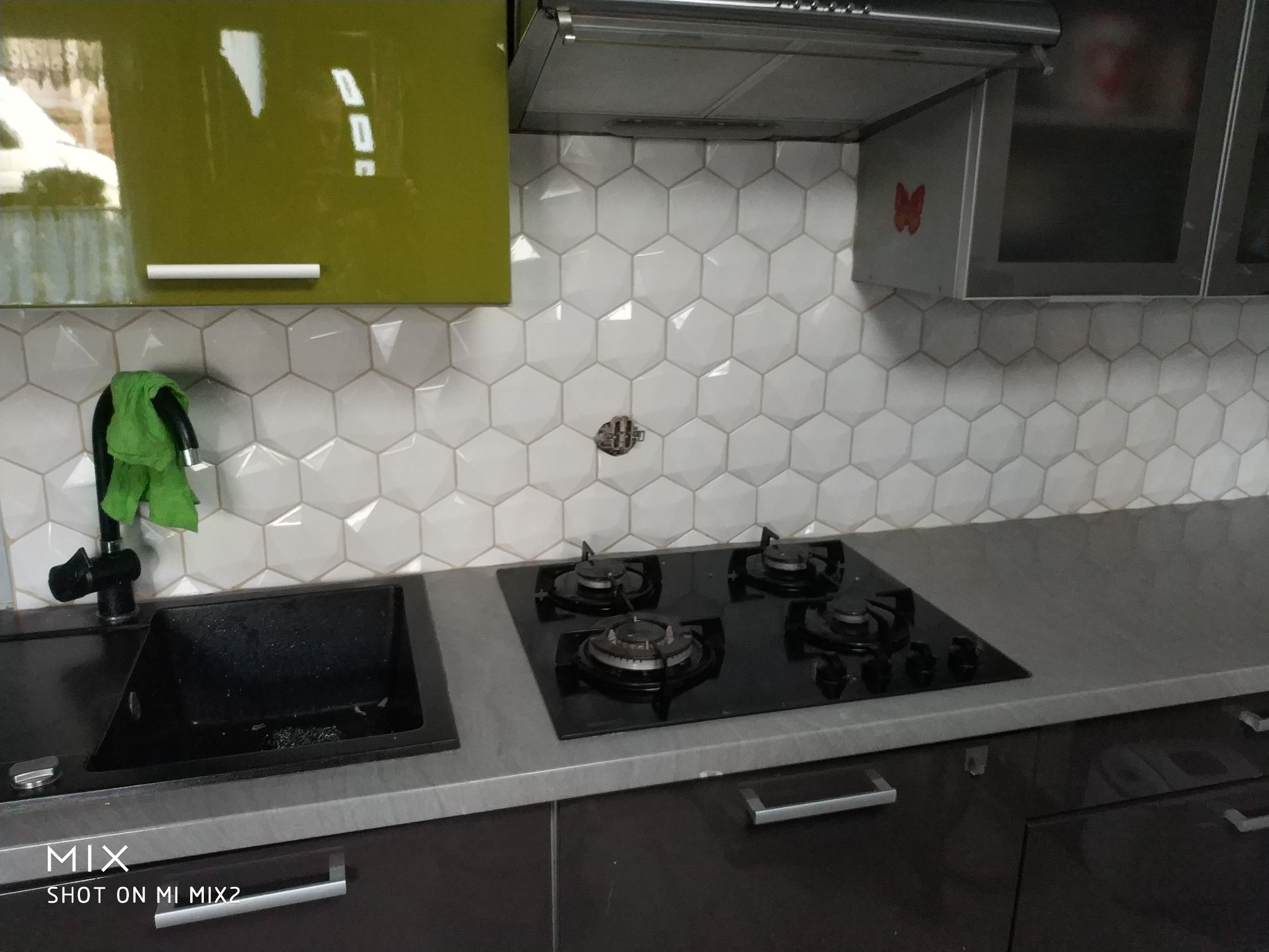 Kuchnia po remoncie: białe heksagonalne płytki na ścianie nad czarnym zlewem i płytą gazową, zielona szafka i okap z naklejką motyla.