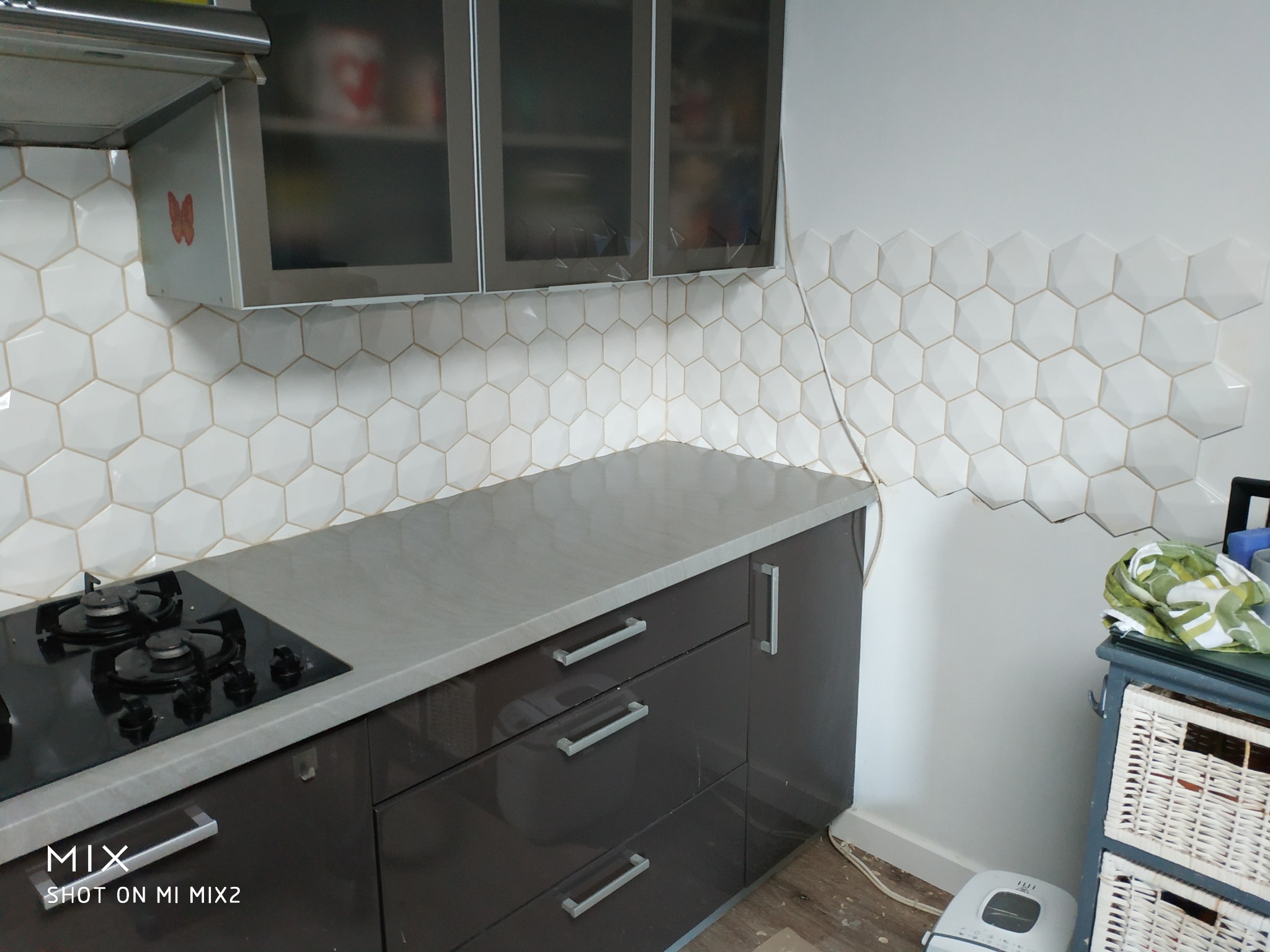 Fragment kuchni z heksagonalnymi płytkami ściennymi w odcieniach bieli, szarym blatem i ciemnymi szafkami, nad którymi wiszą szafki z metalowymi ramami i matowym szkłem.