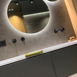 Patrycja Wójcik - Nowoczesna łazienka z okrągłym lustrem podświetlanym LED, szarą umywalką z betonu architektonicznego i czarną armaturą, uzupełniona zieloną ściereczką.