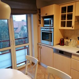 Patrycja Wójcik - Jasna kuchnia z drewnianymi szafkami, sprzętami AGD (piekarnik, mikrofalówka, zmywarka), widok na zewnątrz przez duże okno balkonowe, białe krzesła przy okrągłym stole.