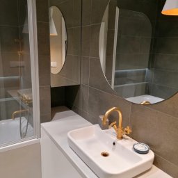 Patrycja Wójcik - Nowoczesna łazienka z szarymi płytkami, białą umywalką i złotą armaturą, odbicie w okrągłym lustrze ukazuje kabinę prysznicową.