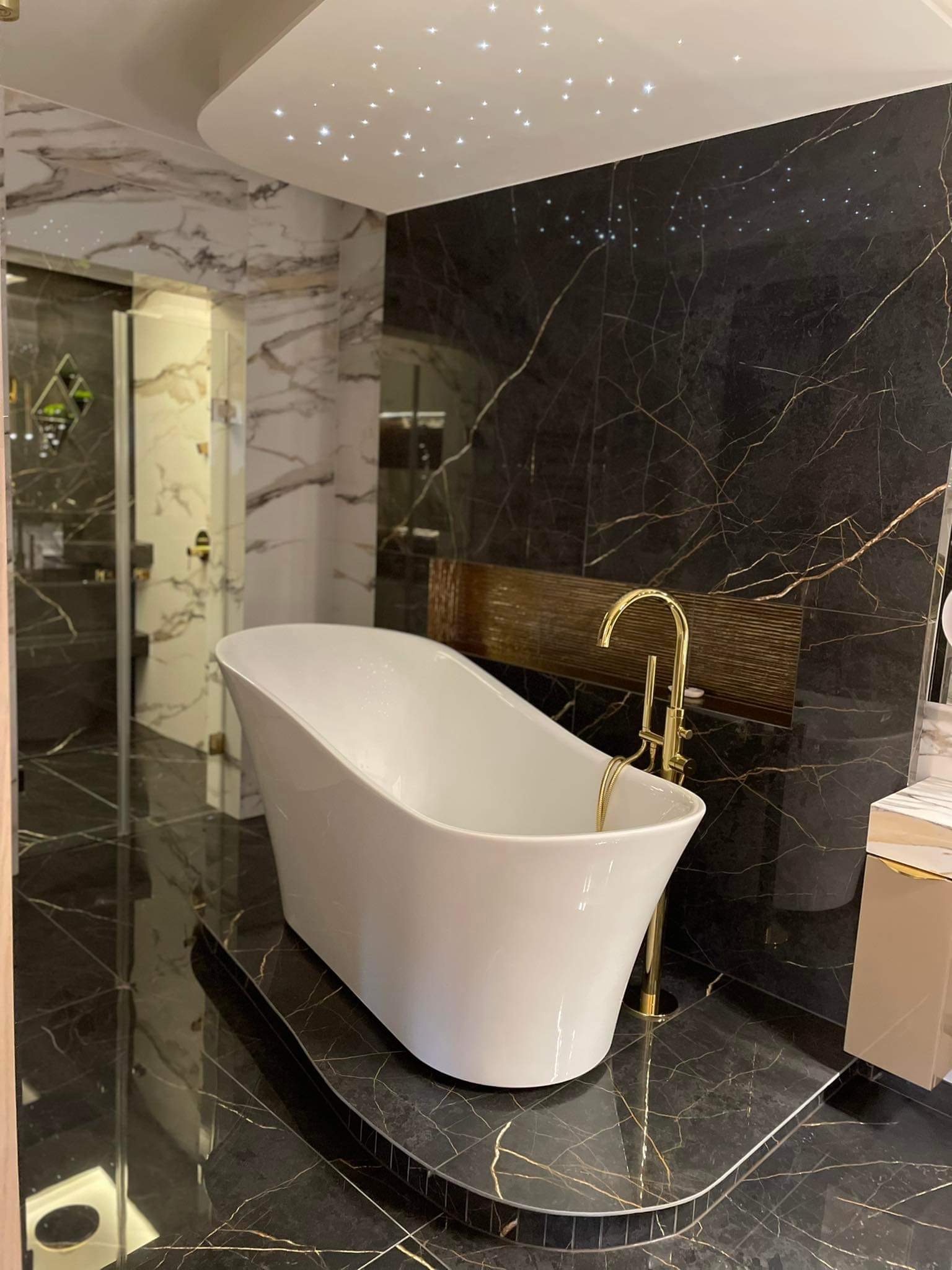 Nowoczesna łazienka z wolnostojącą białą wanną, złotą armaturą i czarnymi marmurowymi ścianami z efektem gwieździstego nieba na suficie.