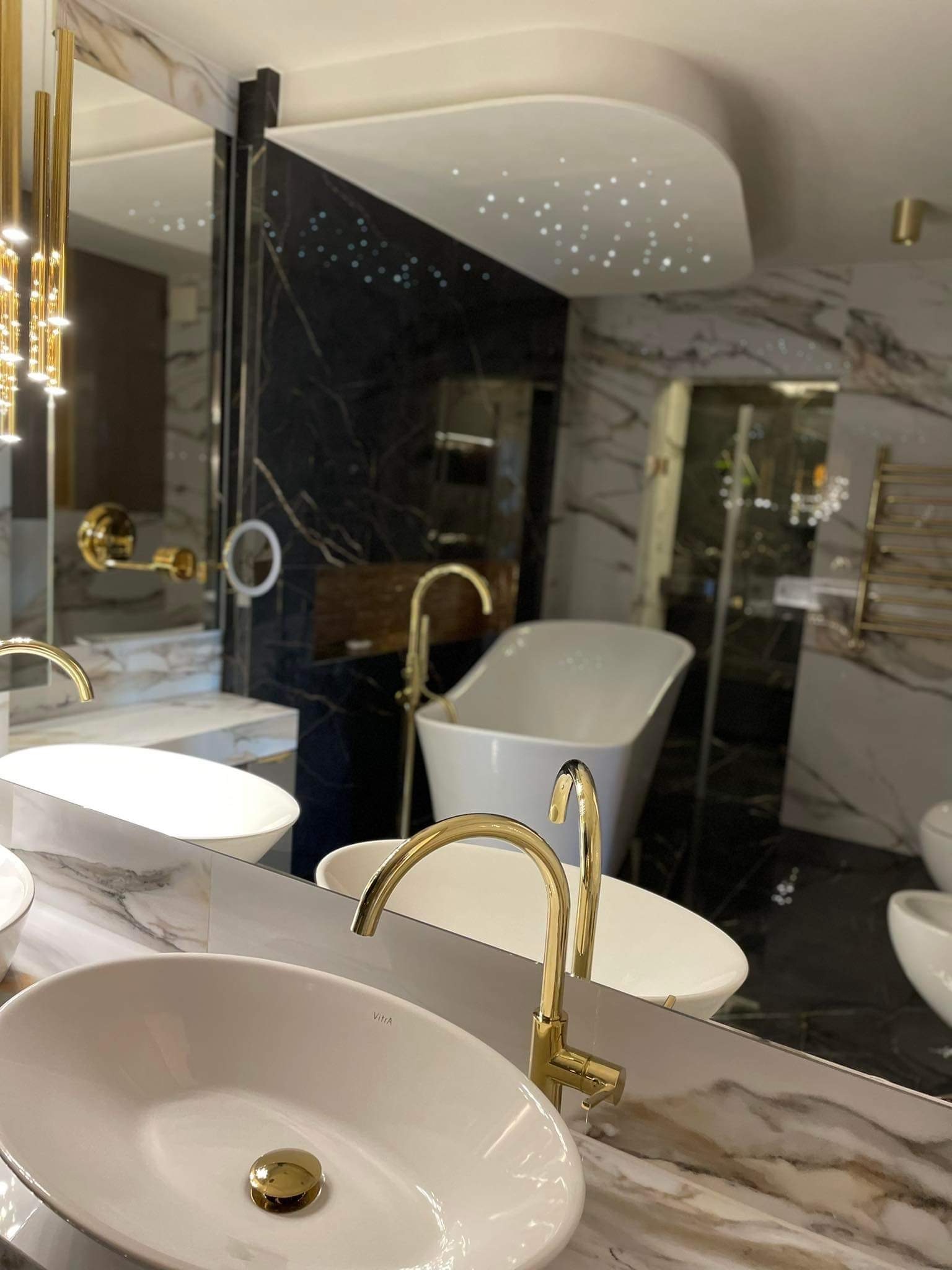 Luksusowa łazienka z marmurowymi ścianami, owalnymi umywalkami z pozłacanymi kranami i wanną wolnostojącą, oświetlona lampą z kryształkami i podświetlanym sufitem.