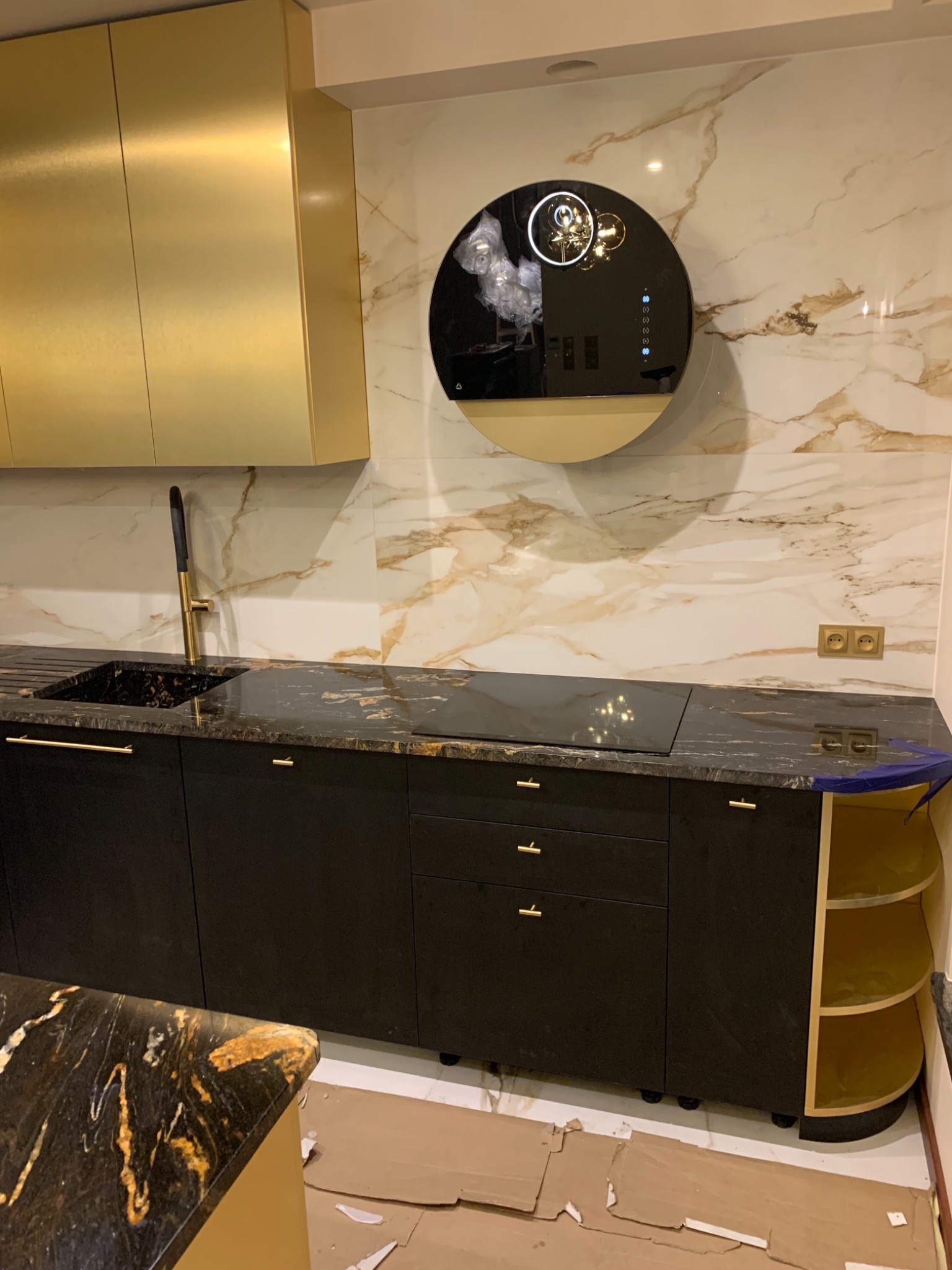 Nowoczesna kuchnia z czarnymi szafkami, złotymi akcentami i okrągłym okapem na tle marmurowej ściany, karton na podłodze sugeruje prace wykończeniowe.