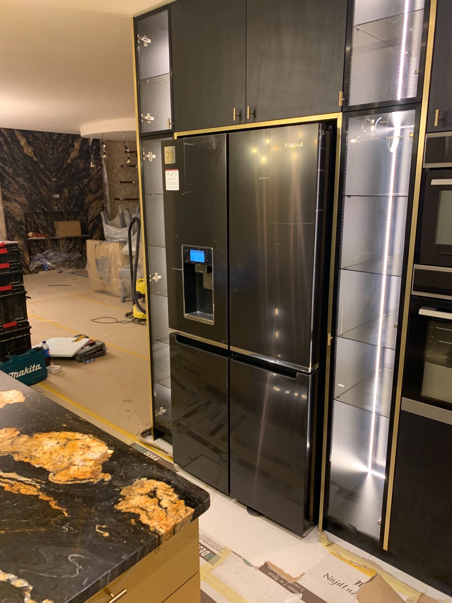 Ciemna zabudowa kuchenna z lodówką Whirlpool, widoczne narzędzia i materiały budowlane na podłodze, sugerujące remont lub wykończenie.