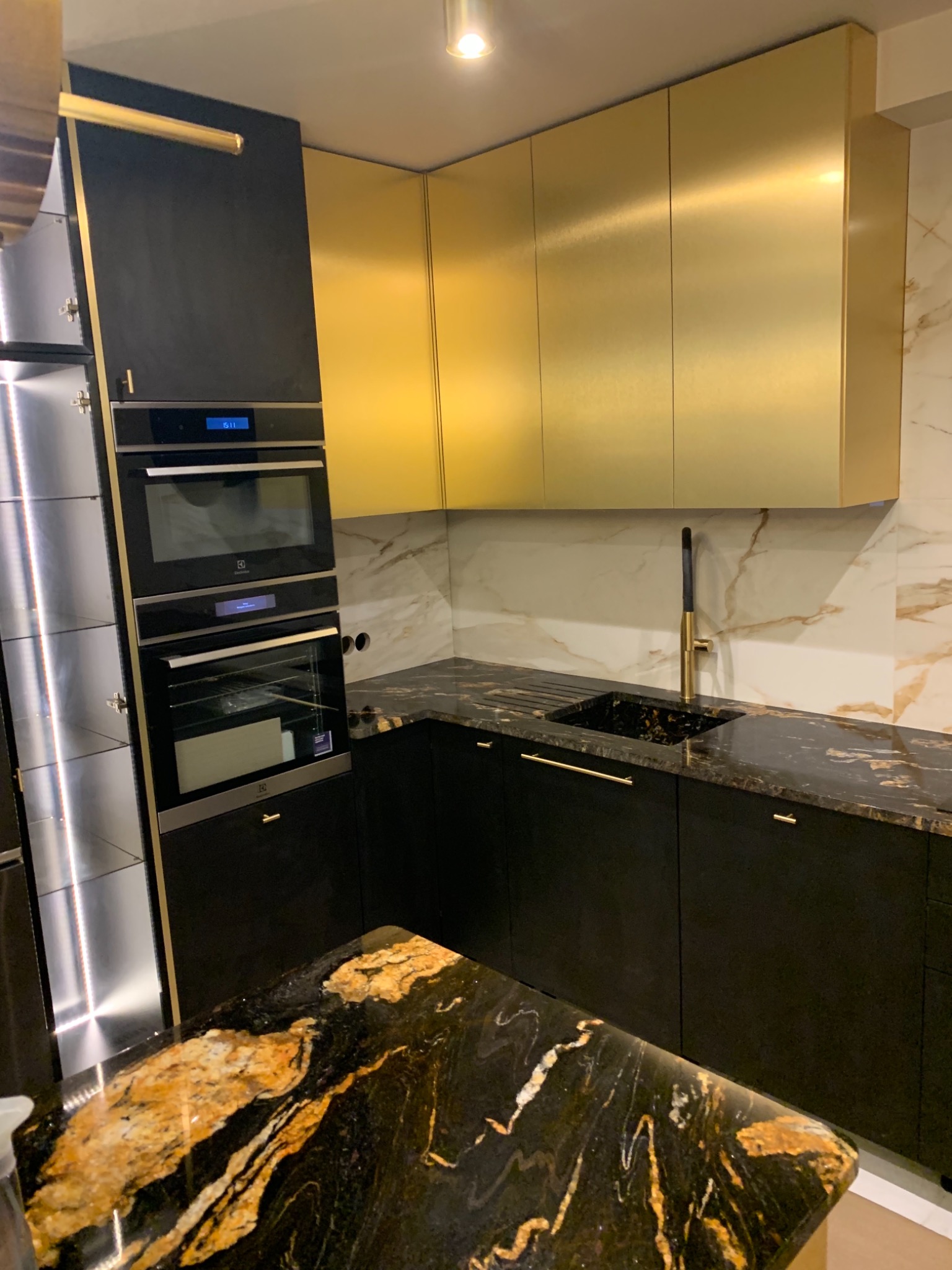 Nowoczesna kuchnia z czarnymi szafkami dolnymi i złotymi górnymi, marmurowy blat i ściana, widoczny piekarnik i mikrofalówka Electrolux, czarny zlew z złotą baterią.