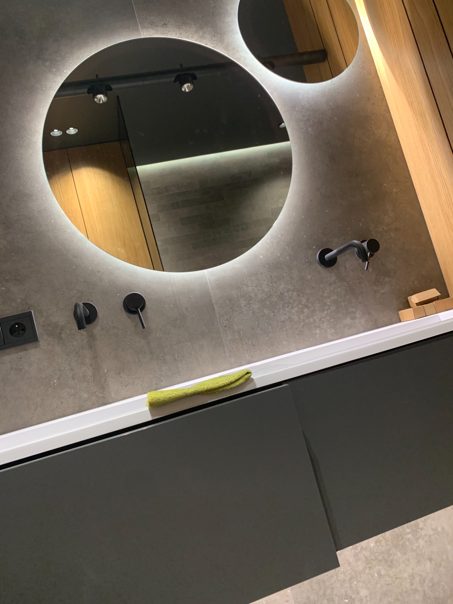 Nowoczesna łazienka z okrągłym lustrem podświetlanym LED, szarą umywalką z betonu architektonicznego i czarną armaturą, uzupełniona zieloną ściereczką.