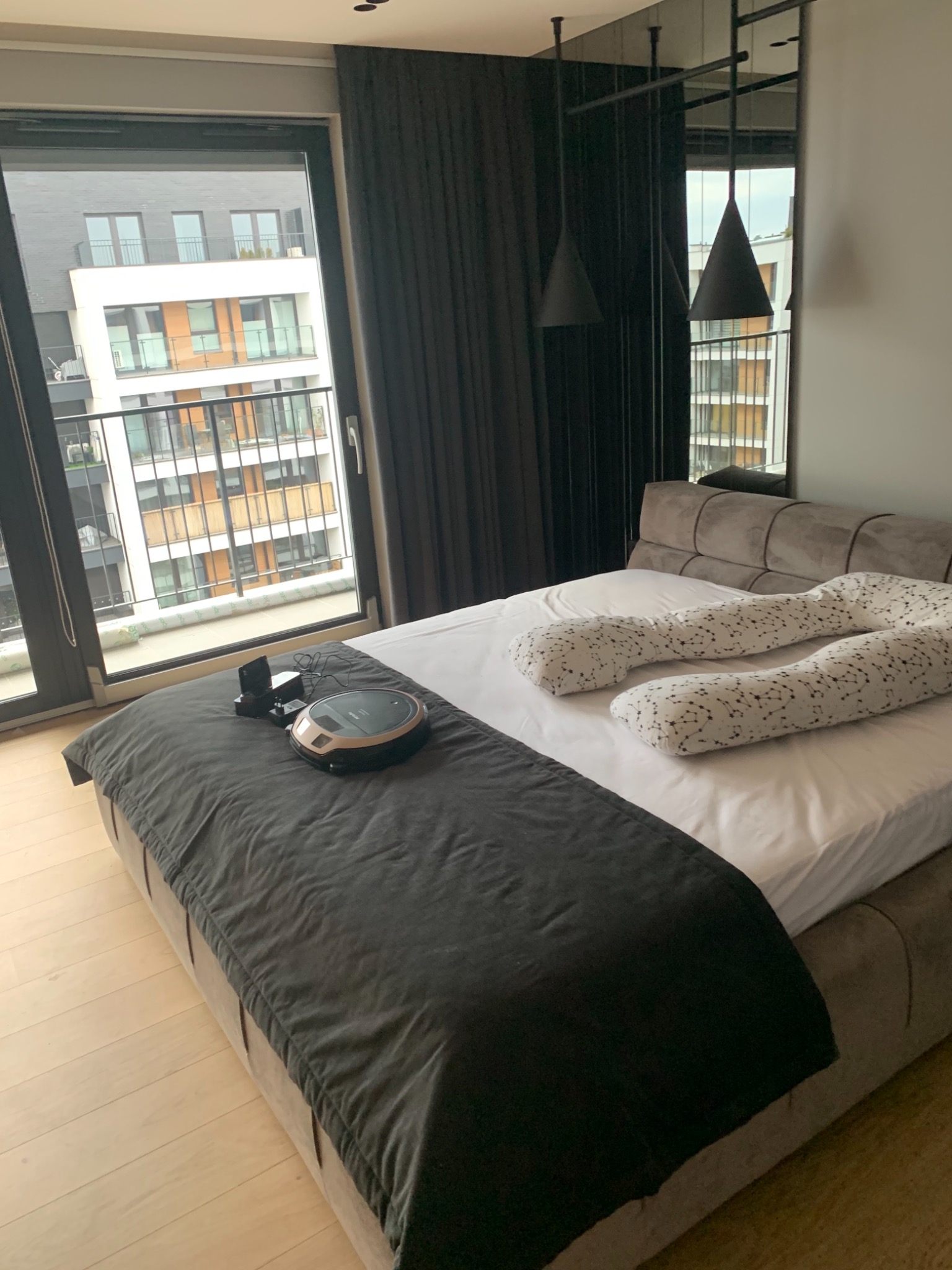 Sypialnia w nowoczesnym apartamencie z łóżkiem, robotem sprzątającym na pościeli, poduszką ciążową w gwiazdki oraz widokiem z balkonu na budynek mieszkalny.