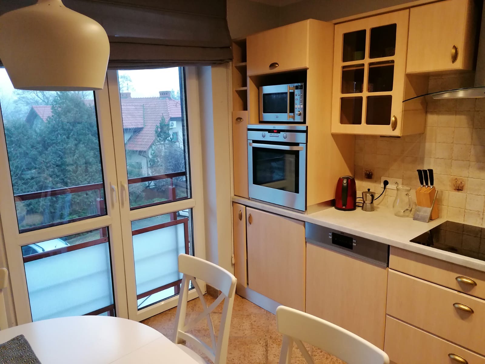 Jasna kuchnia z drewnianymi szafkami, sprzętami AGD (piekarnik, mikrofalówka, zmywarka), widok na zewnątrz przez duże okno balkonowe, białe krzesła przy okrągłym stole.