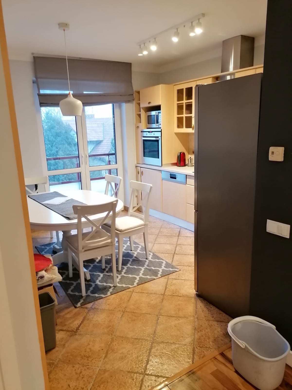 Jasna kuchnia z okrągłym stołem i białymi krzesłami, widoczny fragment podłogi z terakoty i dywanem w geometryczny wzór, obok stoi szary kosz i wiadro.