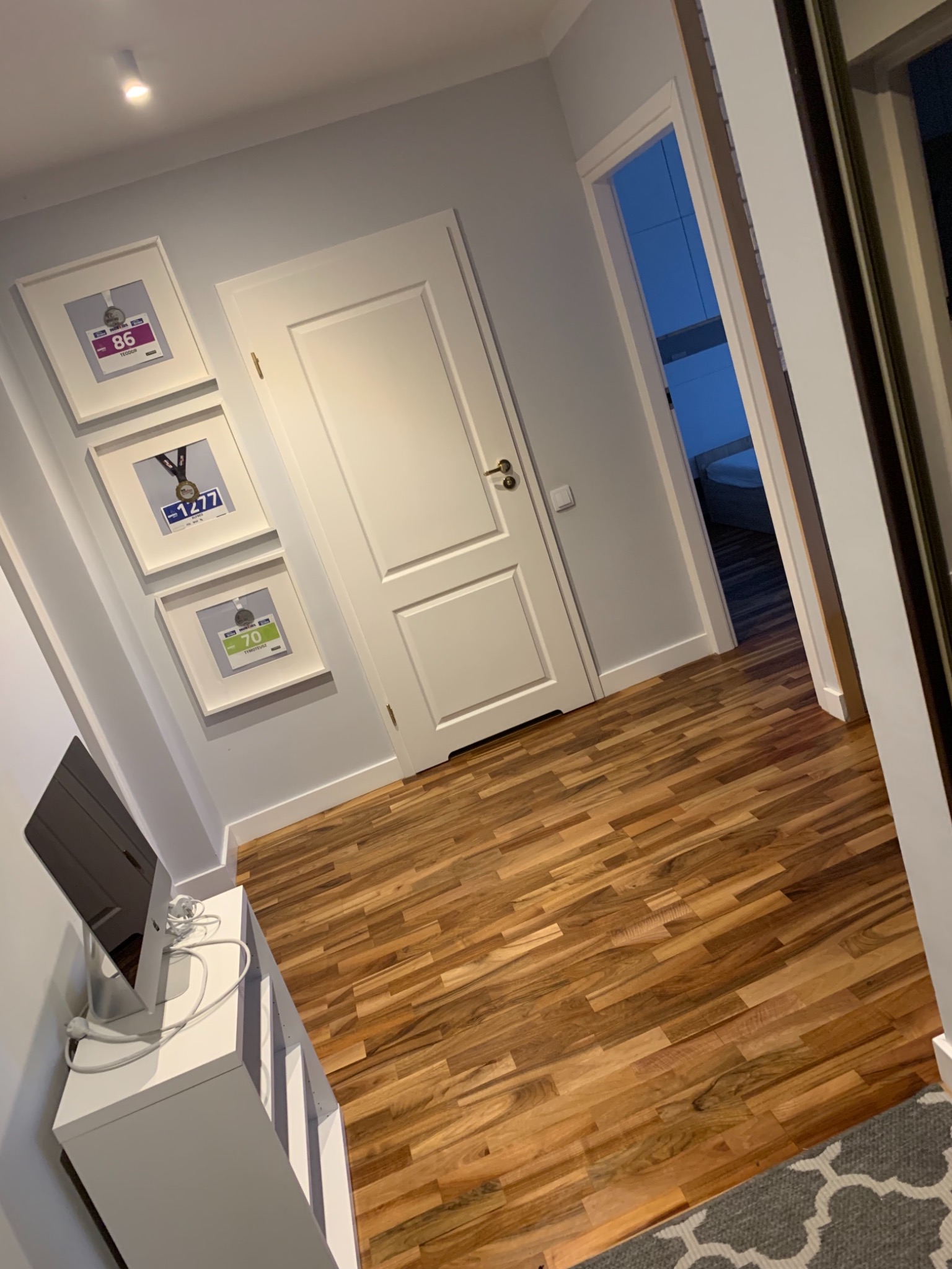 Wnętrze korytarza z drewnianą podłogą, białymi drzwiami i ścianami w odcieniach szarości, na których wiszą oprawione medale sportowe. Widoczny fragment białej szafki z monitorem i kablem...