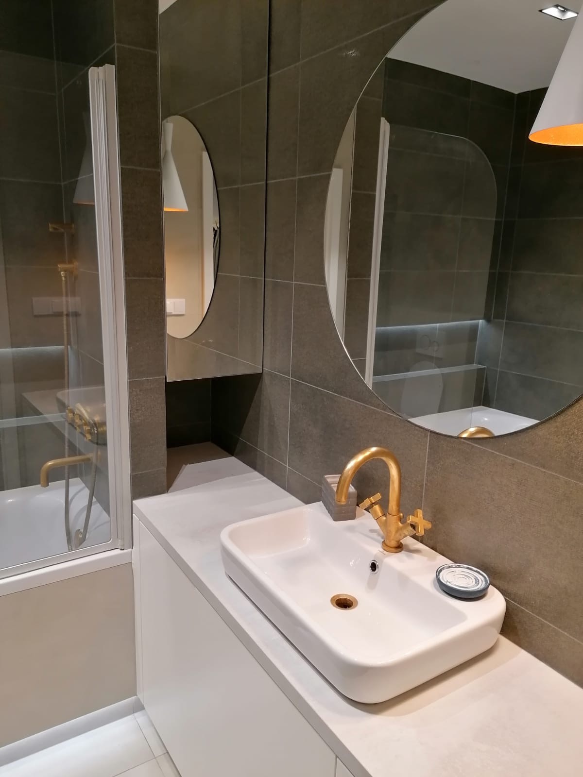 Nowoczesna łazienka z szarymi płytkami, białą umywalką i złotą armaturą, odbicie w okrągłym lustrze ukazuje kabinę prysznicową.