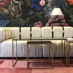 Nowoczesny salon z kremową sofą z paskami, stolikami kawowymi ze złotymi ramami i dywanem w odcieniach różu, na tle ściany w ciemne kwiaty.