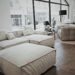 Nowoczesne, modułowe sofy w odcieniach beżu na ciemnej, drewnianej podłodze ułożonej w jodełkę, widok z salonu w Legnicy na ulicę przez duże, łukowe okno.