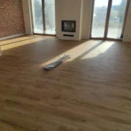 Wnętrze pokoju z nową, jasnobrązową podłogą imitującą drewno, z widoczną stertą paneli na środku, ceglana ściana w tle, duże okna z zasłoniętymi szybami.