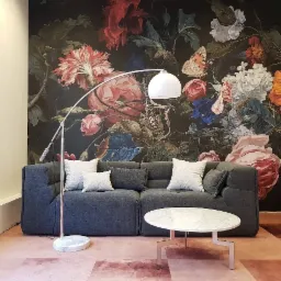 Szary narożnik z poduszkami, okrągły stolik z marmurowym blatem i lampa łukowa na tle ściany z motywem kwiatowym.