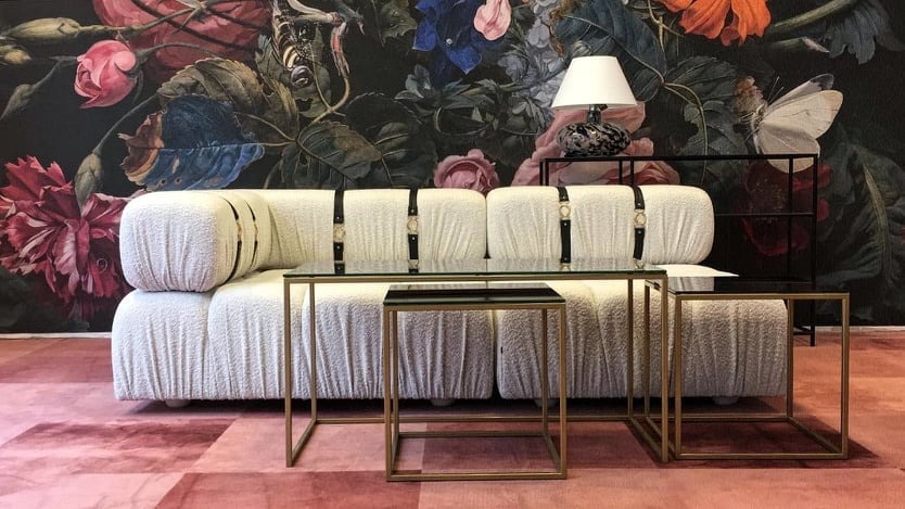 Nowoczesny salon z kremową sofą z paskami, stolikami kawowymi ze złotymi ramami i dywanem w odcieniach różu, na tle ściany w ciemne kwiaty.