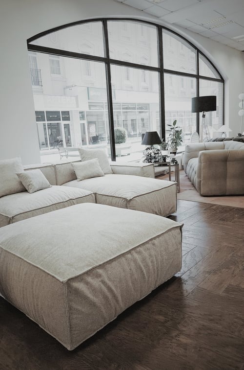 Nowoczesne, modułowe sofy w odcieniach beżu na ciemnej, drewnianej podłodze ułożonej w jodełkę, widok z salonu w Legnicy na ulicę przez duże, łukowe okno.