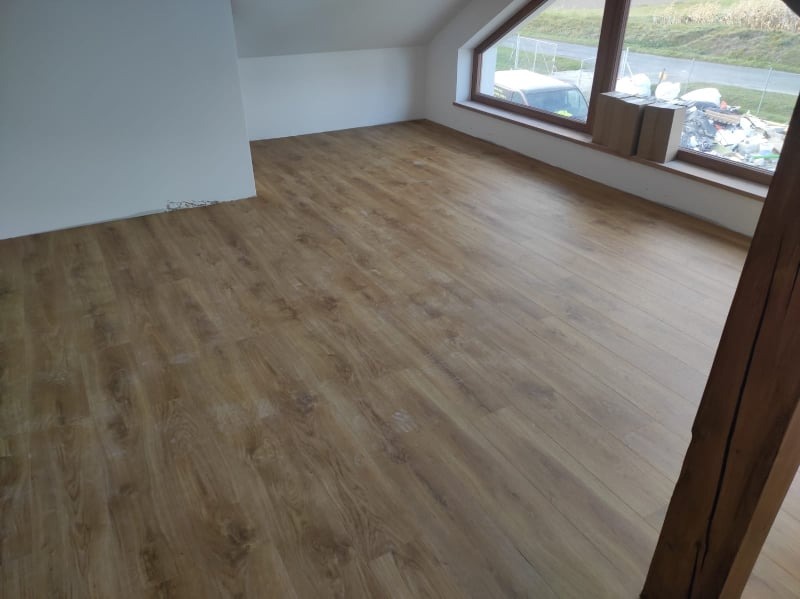 Jasnobrązowa podłoga w nowym pokoju na poddaszu z oknem, widoczne kartony przy oknie i drewniany słup w prawym dolnym rogu.