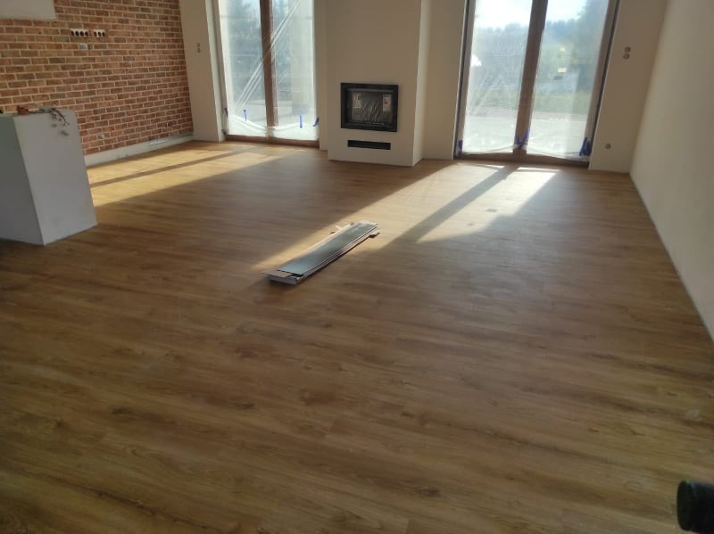 Wnętrze pokoju z nową, jasnobrązową podłogą imitującą drewno, z widoczną stertą paneli na środku, ceglana ściana w tle, duże okna z zasłoniętymi szybami.