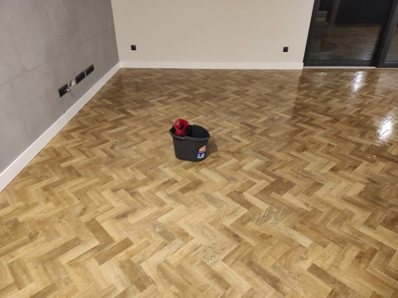 Świeżo położony, lakierowany parkiet w jodełkę, widoczny kubeł z mopem po zakończeniu prac, szara ściana z gniazdkami w tle.
