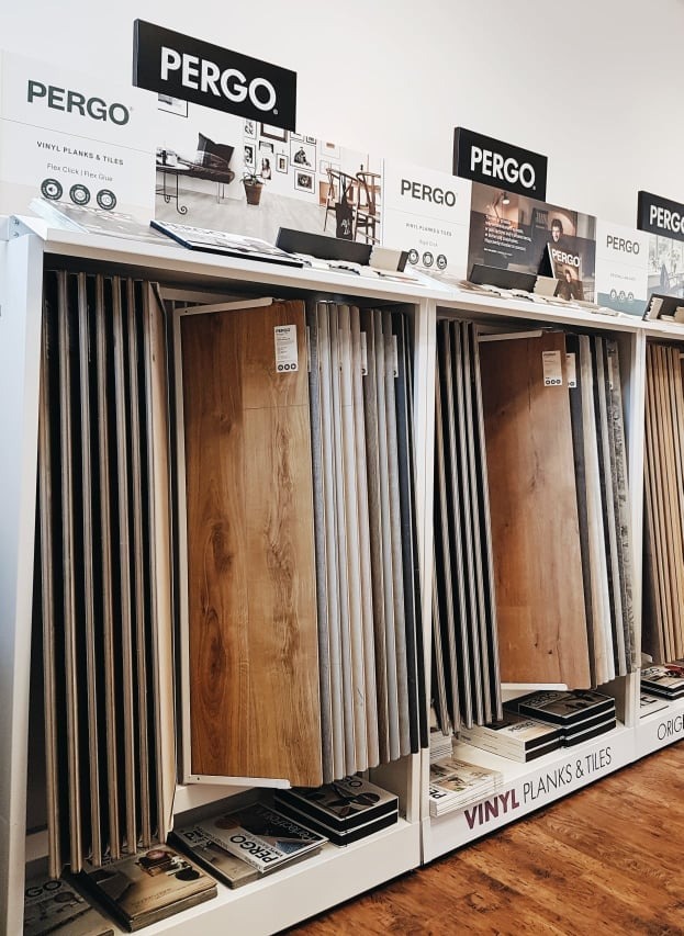 Ekspozycja próbek podłogowych marki Pergo, prezentująca różnorodność wzorów i kolorów paneli winylowych i płytek, umieszczonych na białym regale w sklepie.