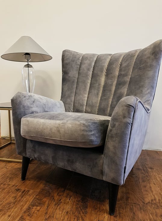 Szare, welurowe krzesło typu uszak z pionowymi przeszyciami na oparciu, stojące na drewnianej podłodze obok stolika z lampą.