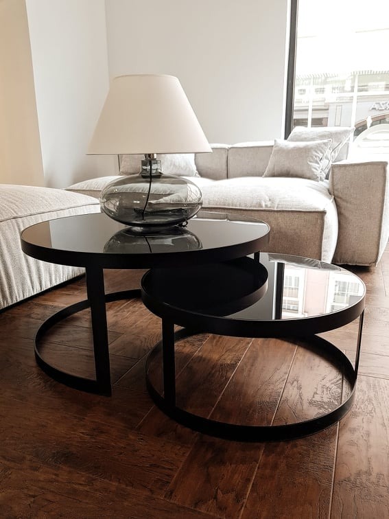 Dwa okrągłe stoliki kawowe z czarnymi ramami i szklanymi blatami na ciemnobrązowej podłodze z paneli, obok jasnej sofy i lampy z szarym szklanym korpusem.