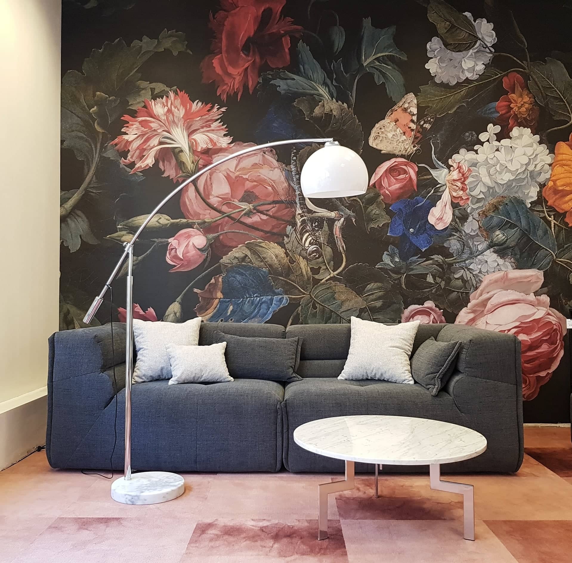 Szary narożnik z poduszkami, okrągły stolik z marmurowym blatem i lampa łukowa na tle ściany z motywem kwiatowym.