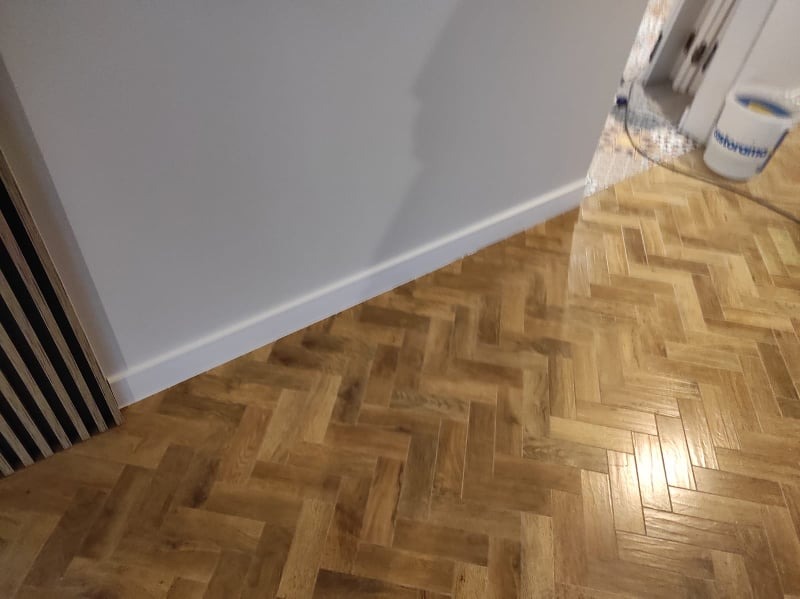 Ukończona podłoga z parkietu ułożonego w jodełkę, widoczna biała listwa przypodłogowa i fragment ściany, w tle wiadro po farbie i fragment drzwi.