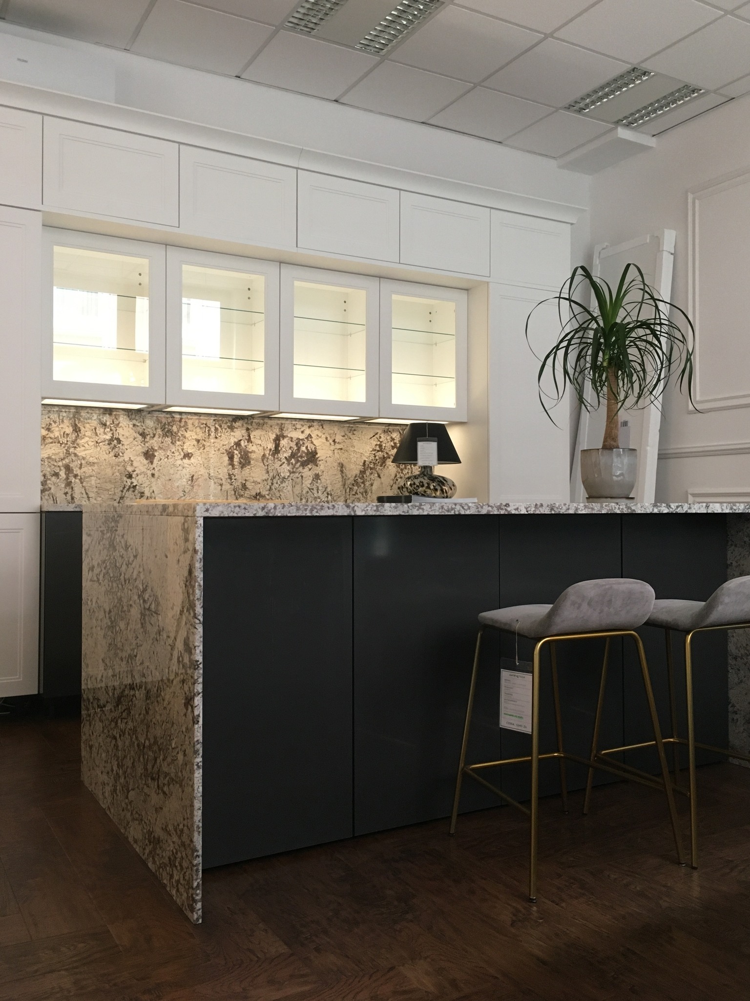 Nowoczesna kuchnia z wyspą wykończoną kamiennym blatem, podświetlane szafki wiszące i drewniana podłoga ułożona w jodełkę.