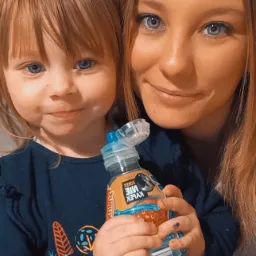 Uśmiechnięta dziewczynka z niebieskimi oczami trzyma w rączkach butelkę wody Kubuś, obok stoi kobieta z podobnymi oczami i włosami w odcieniach blondu.