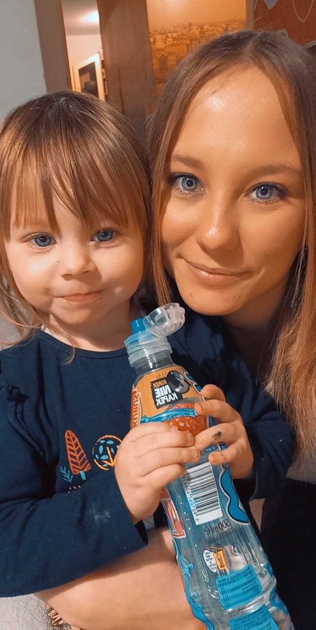 Uśmiechnięta dziewczynka z niebieskimi oczami trzyma w rączkach butelkę wody Kubuś, obok stoi kobieta z podobnymi oczami i włosami w odcieniach blondu.