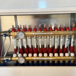 Rozdzielacz ogrzewania podłogowego w szafce, widoczne rury zasilające i powrotne, manometry, etykiety z nazwami pomieszczeń (Salon, Kuchnia, Łazienka, Spiżarnia, Kotłownia, Garaż), oraz zawory...