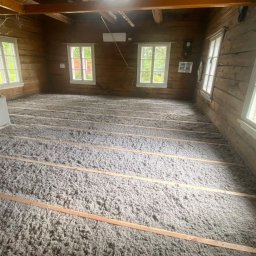 FIRMA HANDLOWO-USŁUGOWA Bartłomiej Biernat - Poddasze starego drewnianego domu w trakcie prac izolacyjnych, widoczne legary i wypełnienie wełną mineralną, trzy okna z białymi ramami w ścianie z bali.