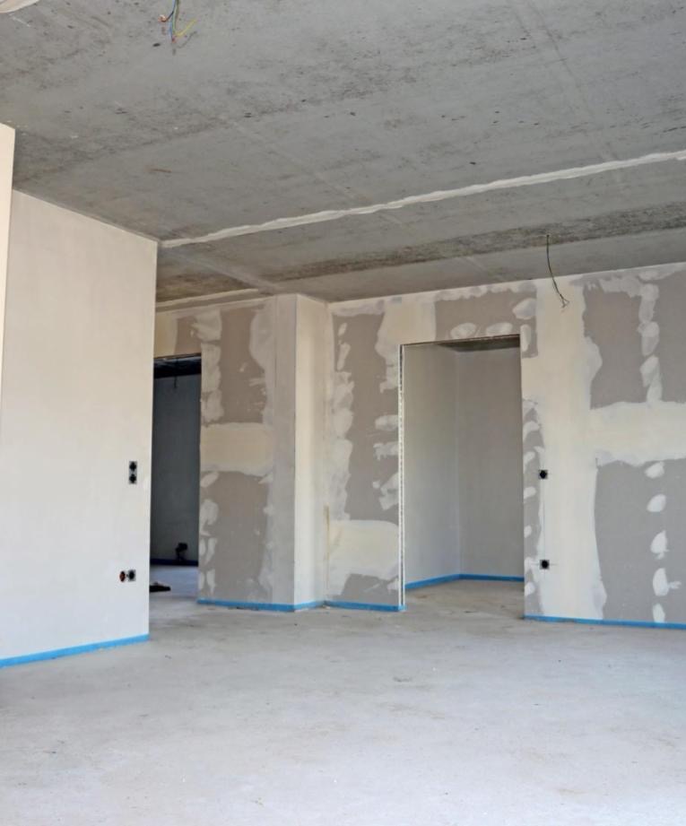 Wnętrze w trakcie prac wykończeniowych: surowy betonowy sufit, ściany częściowo pokryte gładzią gipsową, zabezpieczone niebieską taśmą, widoczne otwory drzwiowe i instalacja elektryczna.