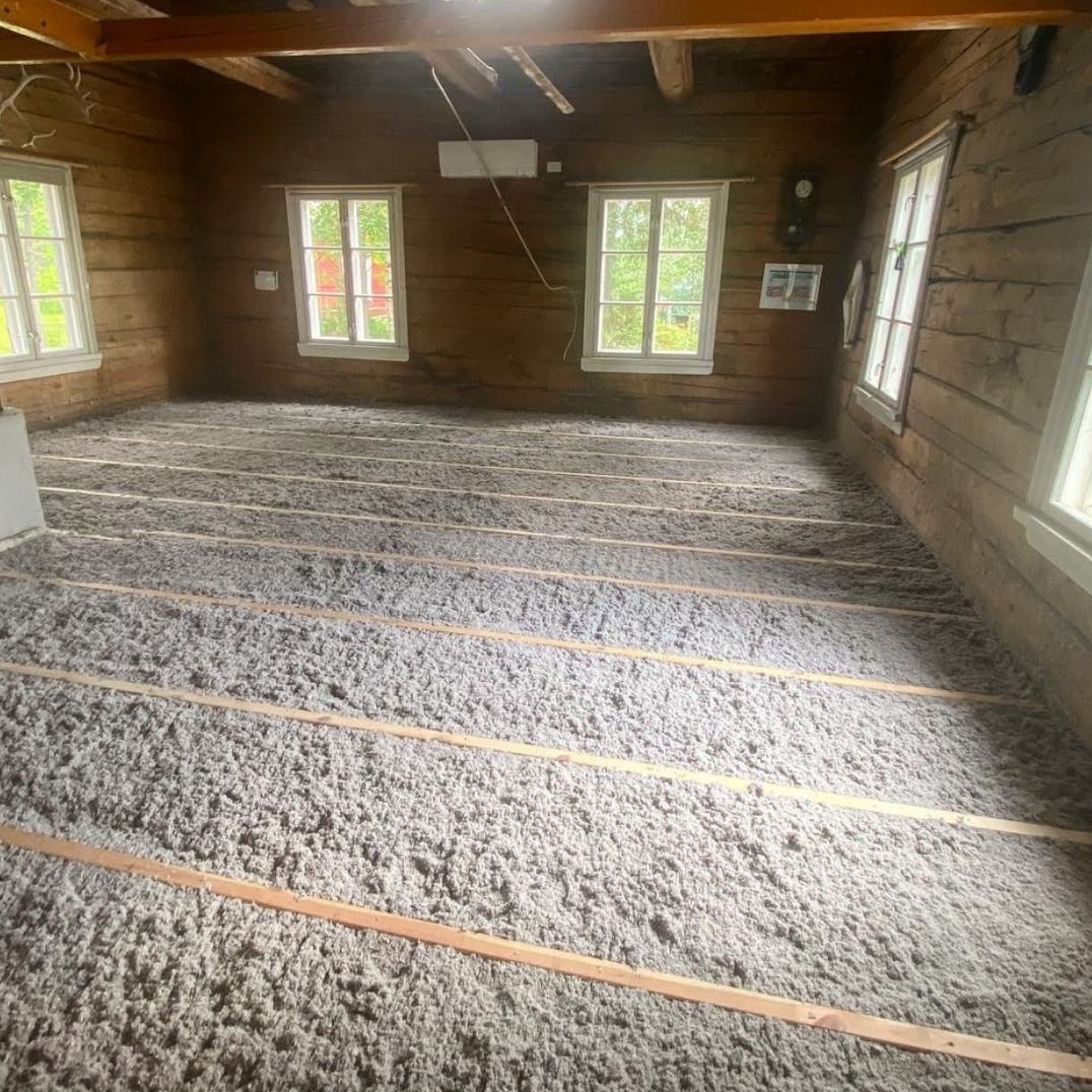 Poddasze starego drewnianego domu w trakcie prac izolacyjnych, widoczne legary i wypełnienie wełną mineralną, trzy okna z białymi ramami w ścianie z bali.