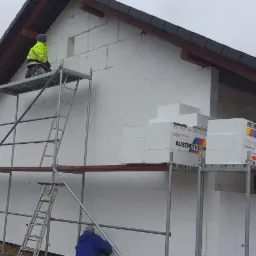 Pracownik w jaskrawej kurtce montuje białe płyty izolacyjne Austrotherm na elewacji domu jednorodzinnego ze szkieletem z pustaków, widoczne rusztowanie i zapas materiału na podeście, szara pogoda.