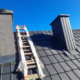 Szary dach z dach&oacute;wki ceramicznej z kominem obłożonym tynkiem mozaikowym i obr&oacute;bką blacharską, drabina oparta o komin zabezpieczona folią malarską, w tle błękitne niebo.