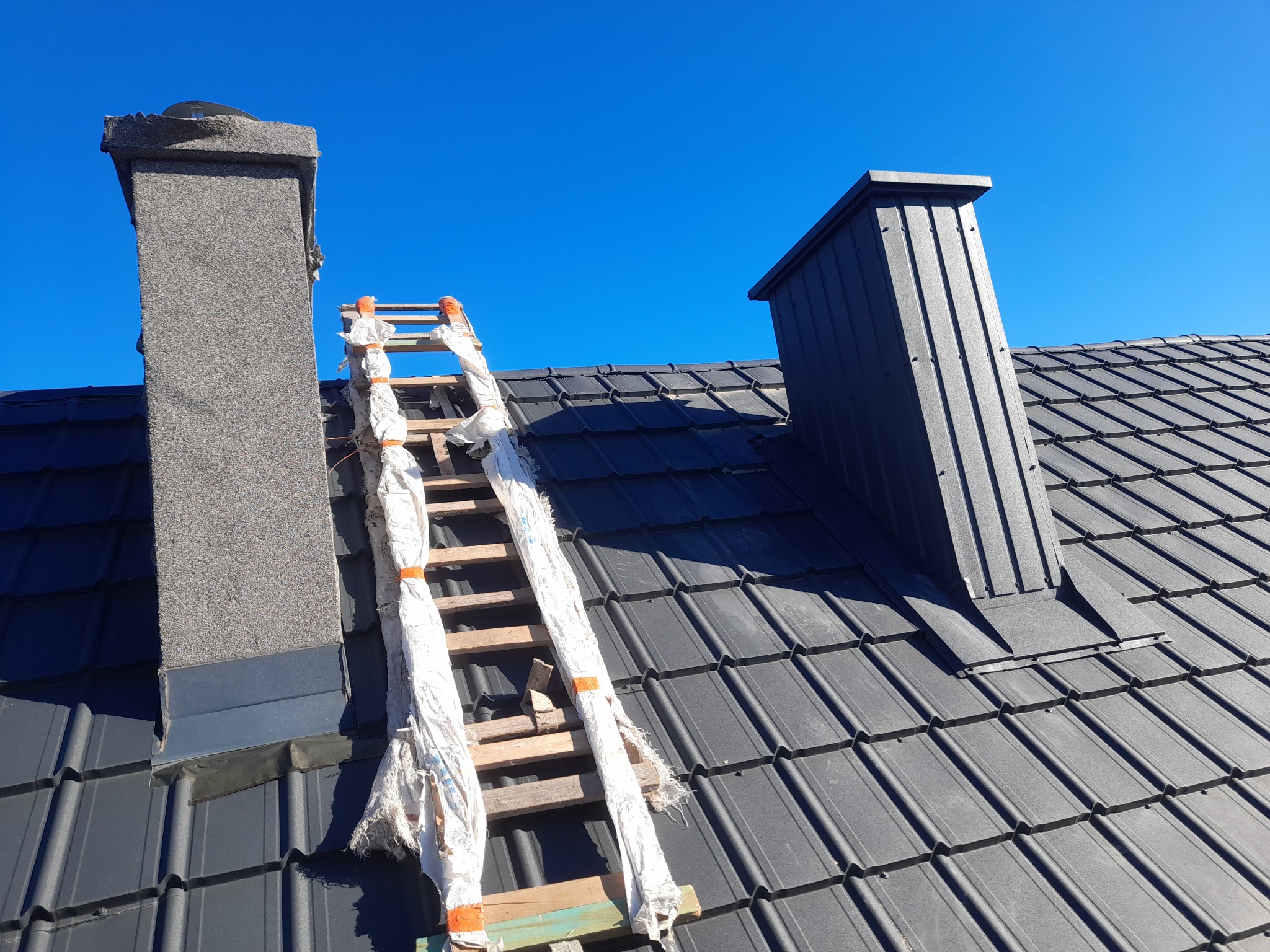 Szary dach z dachówki ceramicznej z kominem obłożonym tynkiem mozaikowym i obróbką blacharską, drabina oparta o komin zabezpieczona folią malarską, w tle błękitne niebo.