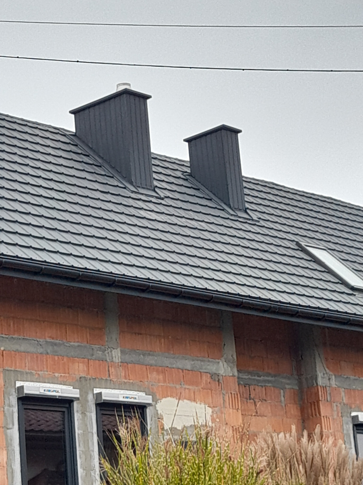 Szary dach z dwoma kominami, okno dachowe, ściany z czerwonej cegły w trakcie budowy, widoczne okna z roletami zewnętrznymi.