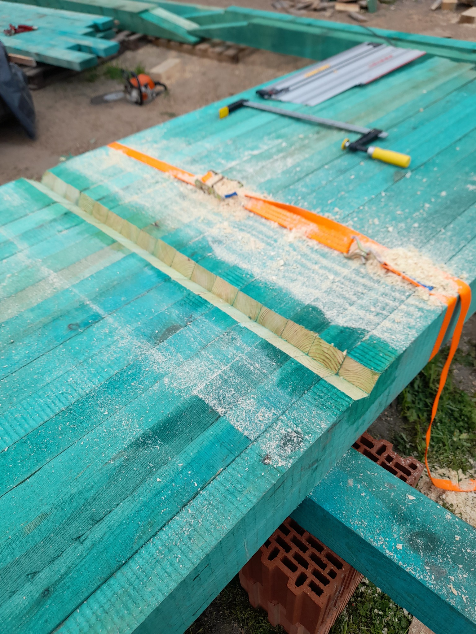 Przygotowane, pomalowane na turkusowo elementy drewniane konstrukcji, spięte pomarańczowym pasem transportowym i zabezpieczone ściskiem stolarskim, leżące na cegłach, z widocznymi trocinami po...