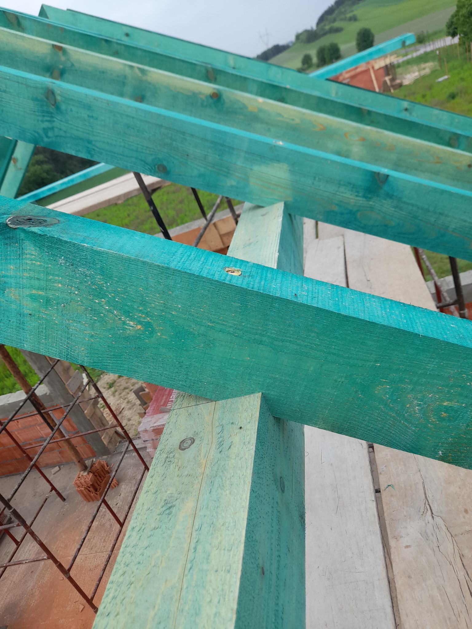 Elementy więźby dachowej, pomalowane na turkusowo, widoczne z dołu, z fragmentami deskowania i rusztowania w tle.
