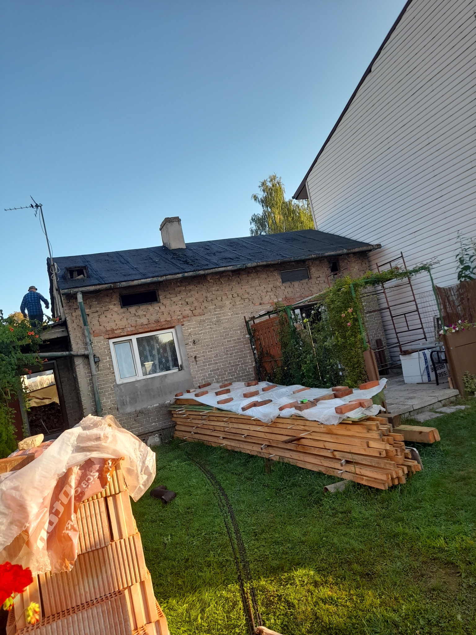 Prace dekarskie przy starym, ceglanym domu: dekarz na dachu, przygotowane deski i cegły na trawniku, widoczny komin i antena.