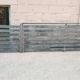 Surowe, niepomalowane przęsła ogrodzenia metalowego z poziomych profili, przygotowane do montażu, składowane przed budynkiem z cegły.