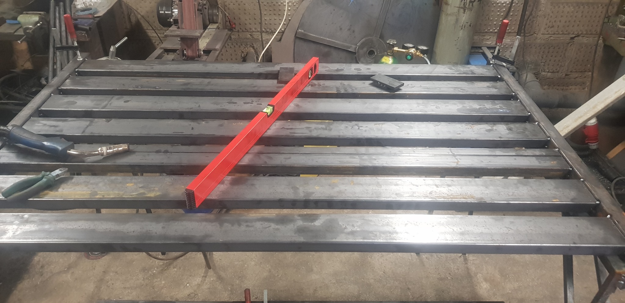 Konstrukcja metalowa w trakcie spawania, widoczna poziomica, spawarka i zaciski, warsztat w tle.