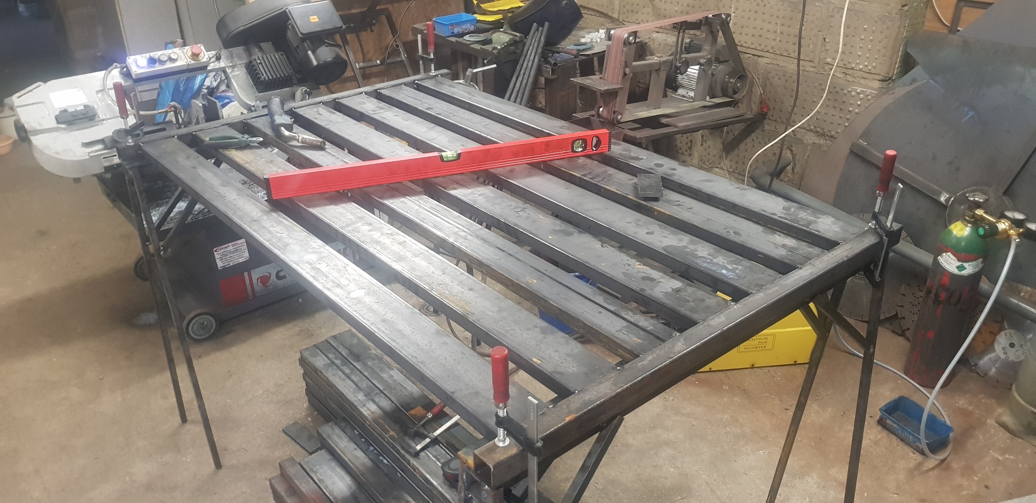 Stół spawalniczy z metalowych profili w trakcie budowy, poziomica spoczywa na konstrukcji, widoczne narzędzia i materiały spawalnicze w warsztacie.