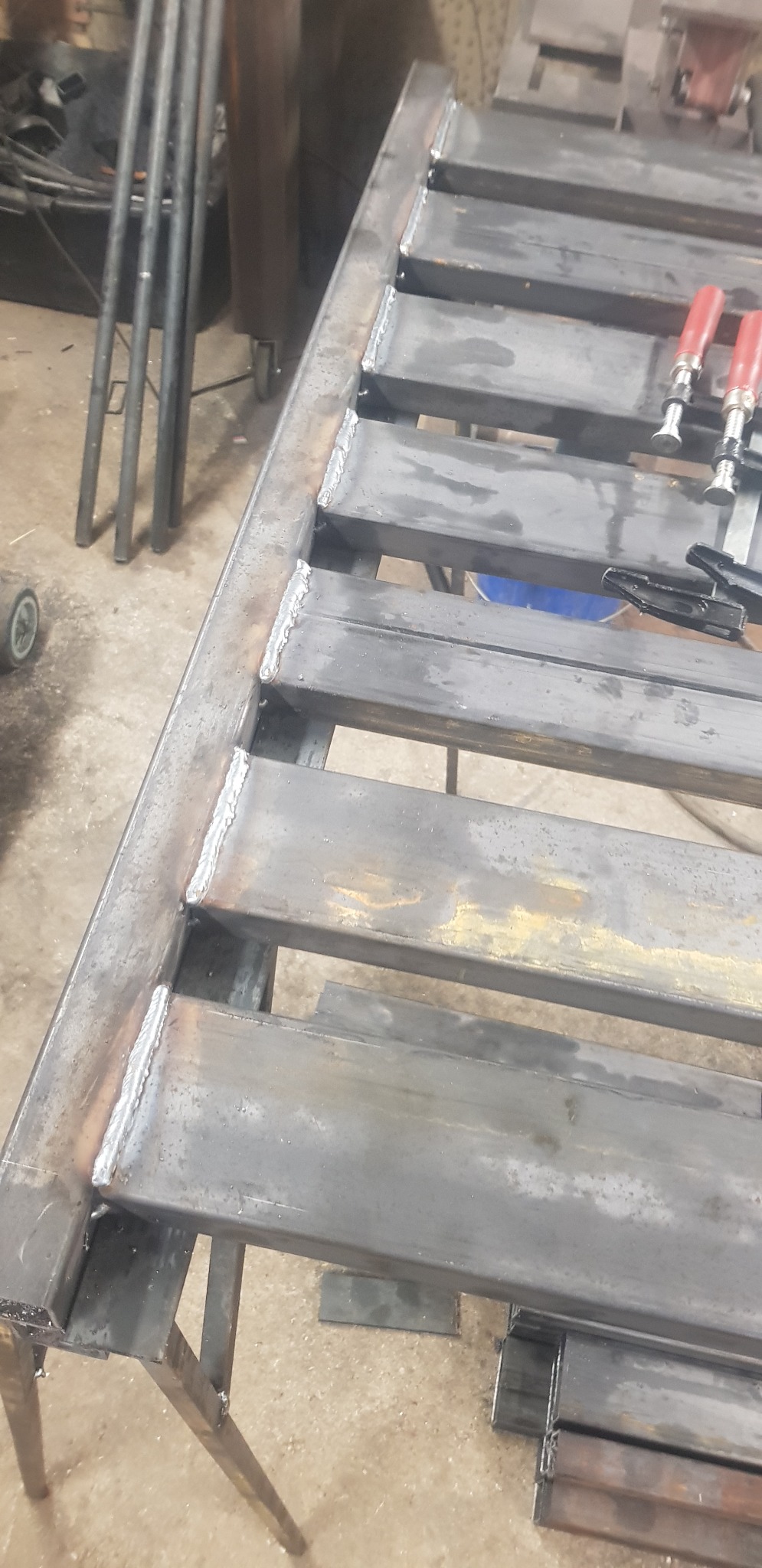Konstrukcja metalowa z przyspawanymi poprzeczkami, częściowo zardzewiała, leżąca na stole warsztatowym, widoczne ślady spawania i narzędzia, w tle rury.