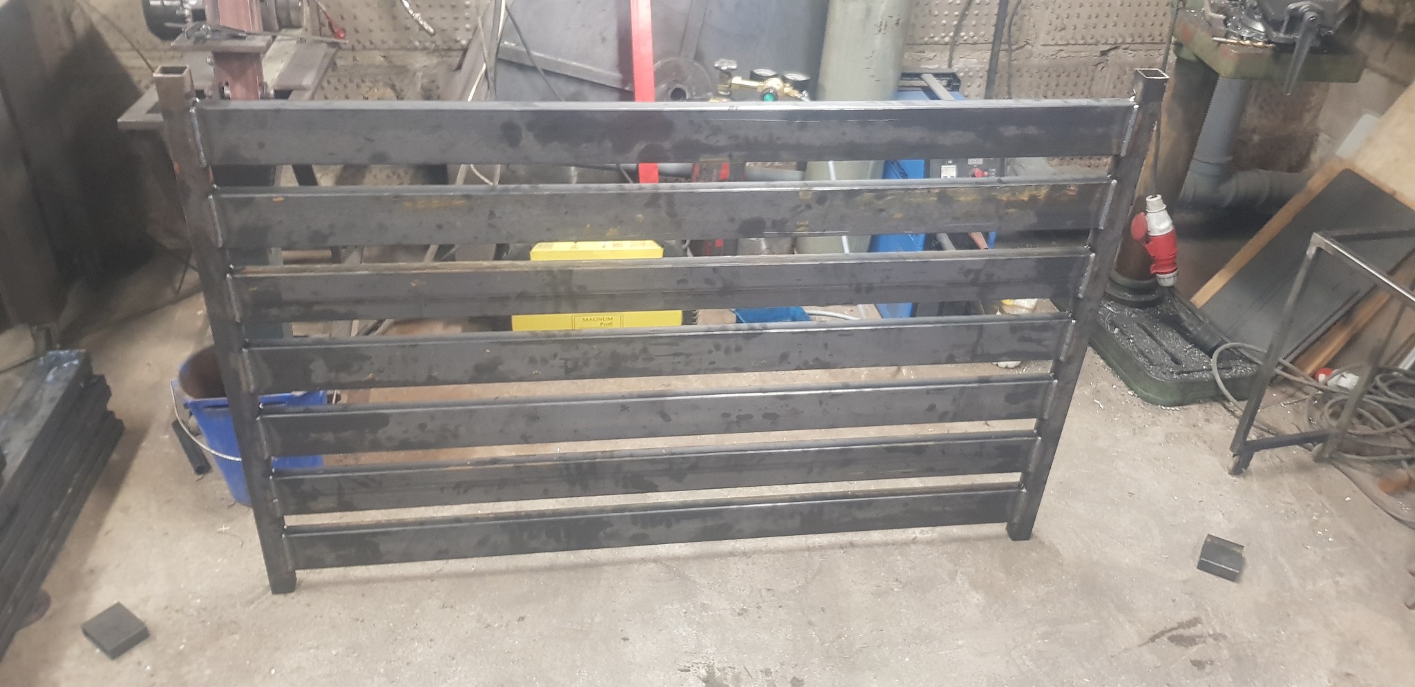 Metalowa konstrukcja z poziomych profili w warsztacie, widoczne ślady spawania i obróbki, nieoszlifowane spawy, w tle narzędzia i maszyny warsztatowe.